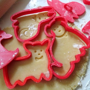 May include: Red plastic cookie cutters in the shapes of a deer, a ghost, a bear, and a snowman on a sheet of unbaked cookie dough.