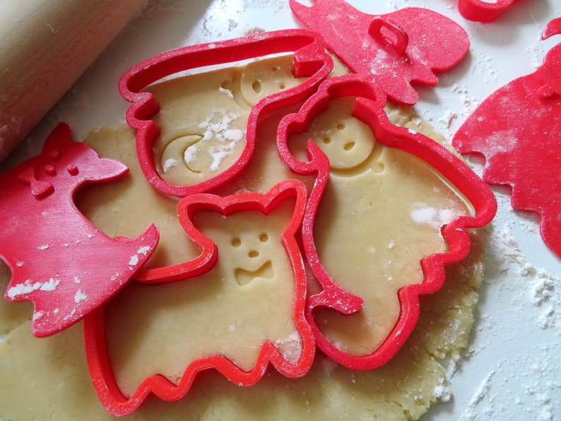 May include: Red plastic cookie cutters in the shapes of a deer, a ghost, a bear, and a snowman on a sheet of unbaked cookie dough.