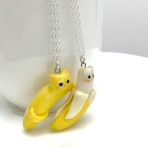 May include: Two necklaces with yellow and white cat-shaped banana pendants. The cats are facing each other and have black eyes.
