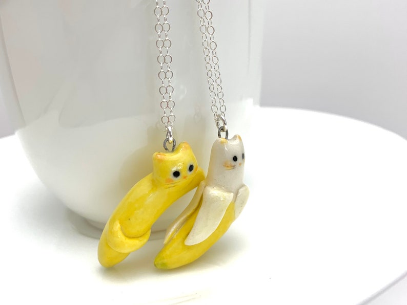 May include: Two necklaces with yellow and white cat-shaped banana pendants. The cats are facing each other and have black eyes.