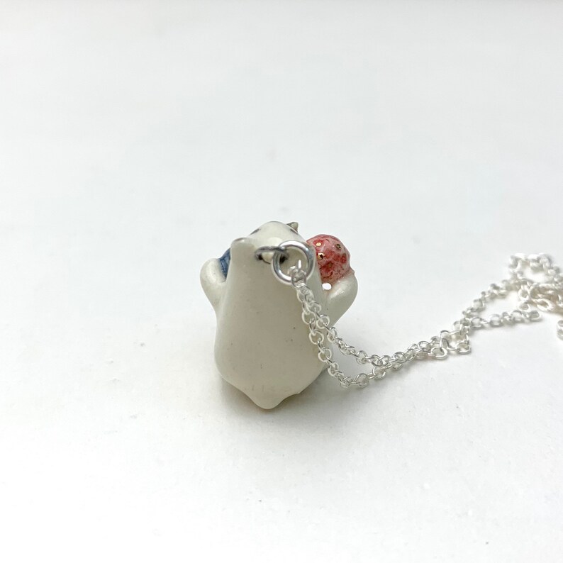 May include: White ceramic pendant necklace with a small red and white ceramic ball. The pendant is shaped like a small animal with a blue and white design. The necklace is silver.