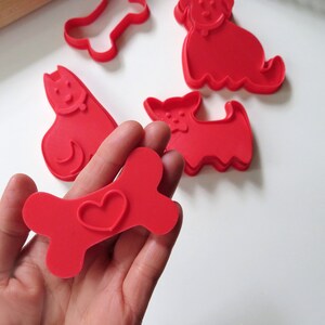 May include: A set of red plastic cookie cutters shaped like a bone with a heart, a dog, and a cat.