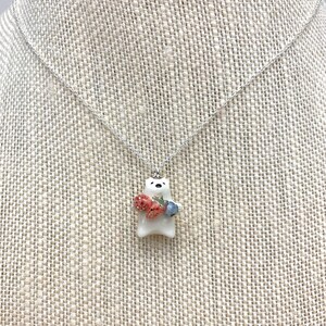 May include: A white ceramic bear pendant necklace with a silver chain. The bear is holding a bunch of red strawberries and blue blueberries.