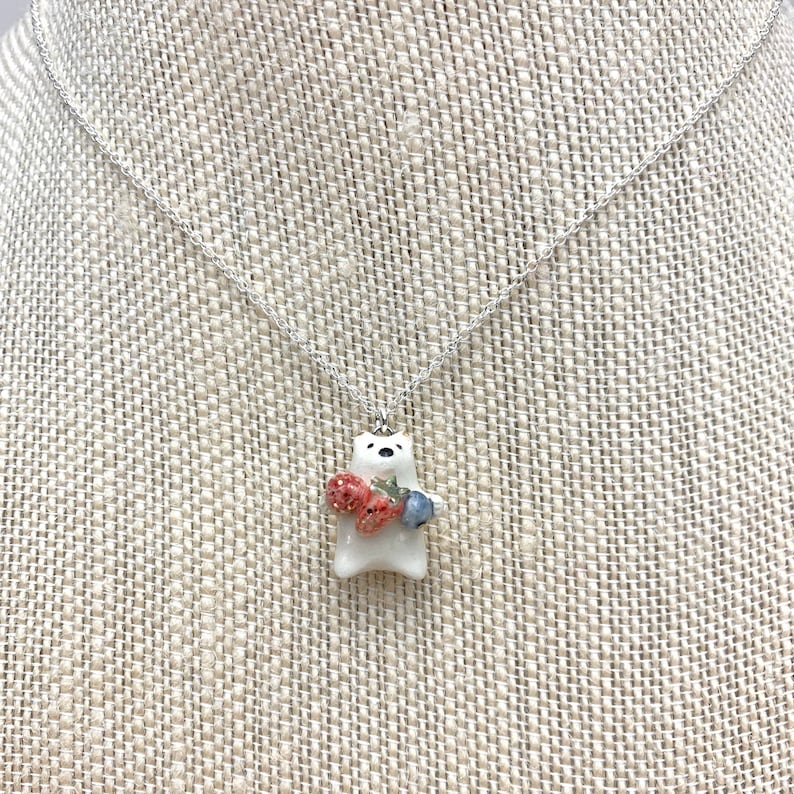 May include: A white ceramic bear pendant necklace with a silver chain. The bear is holding a bunch of red strawberries and blue blueberries.