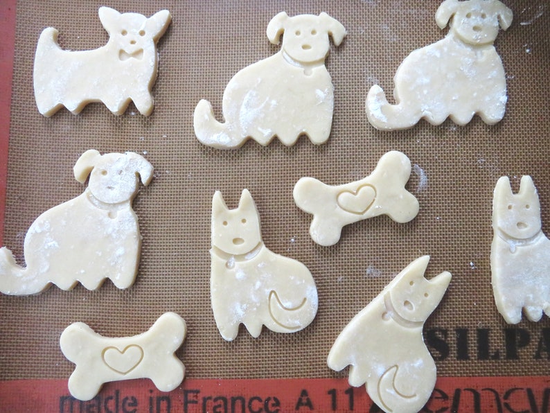 May include: A baking sheet with nine unbaked dog-shaped and bone-shaped sugar cookies. The cookies are dusted with flour and have heart-shaped cutouts.
