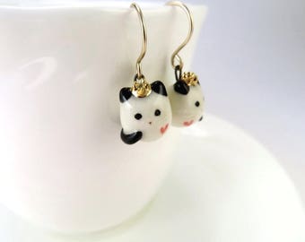 Puede incluir: Un par de pendientes colgantes con un oso panda de cerámica blanca con ojos negros y un corazón rosa en el pecho. Cada panda lleva una corona dorada.