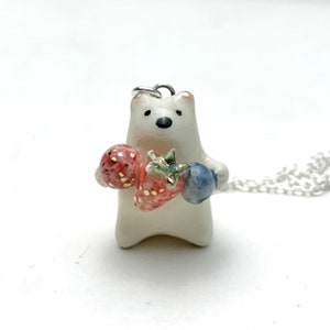 May include: A white ceramic bear pendant necklace with a silver chain. The bear is holding two strawberries and a blueberry.