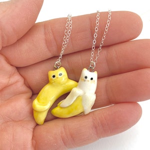 May include: Two silver necklaces with ceramic cat pendants. One cat is yellow and the other is white. Both cats are shaped like bananas.