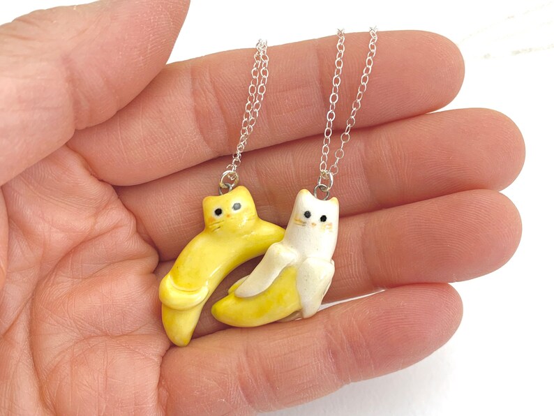 May include: Two silver necklaces with ceramic cat pendants. One cat is yellow and the other is white. Both cats are shaped like bananas.