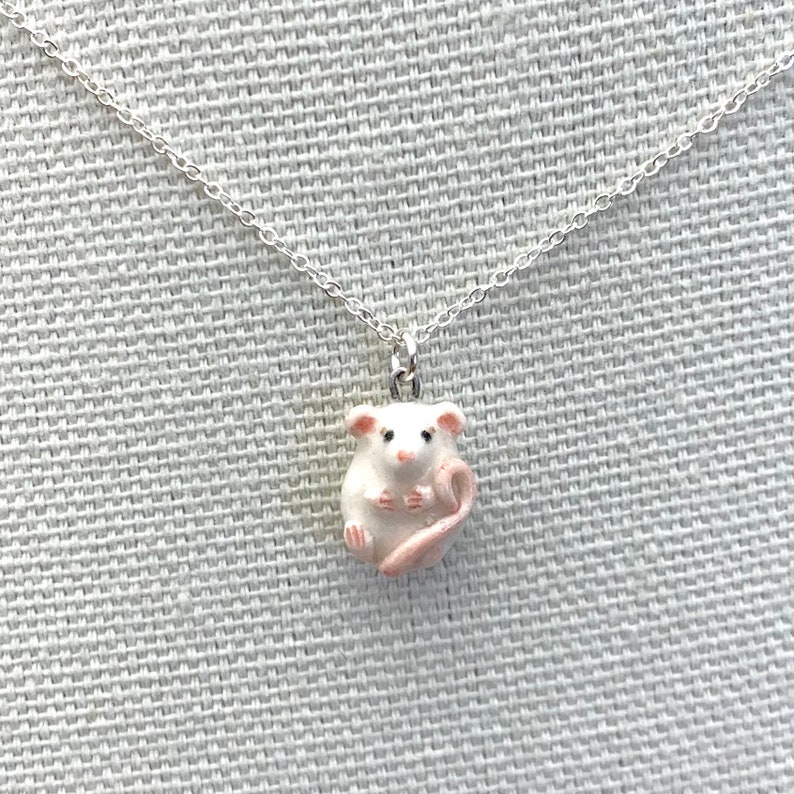 May include: A silver chain necklace with a white ceramic mouse pendant. The mouse has pink ears and a pink tail.