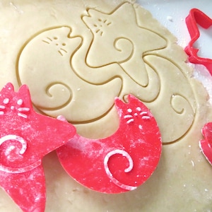 May include: Two red cat-shaped cookie cutters on a sheet of unbaked cookie dough. The cookie cutters are on top of the dough, and the dough is lightly dusted with flour. The cookie cutters are in the shape of a cat with a curled tail.