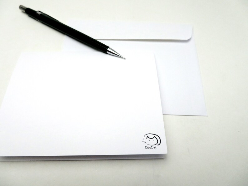 May include: A blank white card with a small black cat logo that says "ChicCraft" in the bottom right corner. A black pencil is lying on top of a white envelope.