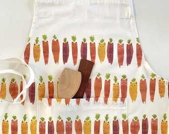 Puede incluir: Delantal de algodón blanco con un patrón repetido de gatos naranja, amarillo y marrón vestidos de zanahorias. El delantal tiene dos bolsillos y una cuchara de madera marrón descansa en uno de los bolsillos.