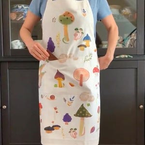 Op de afbeelding: Een witte schort met een kleurrijke paddenstoelprint. De schort heeft een grote zak aan de voorkant en is perfect om te koken of te bakken.