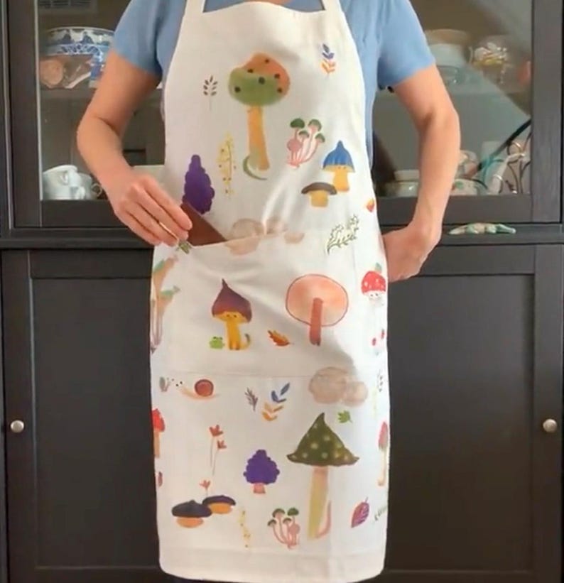 Op de afbeelding: Een witte schort met een kleurrijke paddenstoelprint. De schort heeft een grote zak aan de voorkant en is perfect om te koken of te bakken.