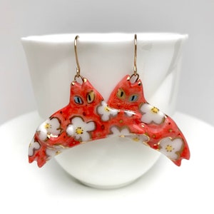 May include: A pair of red cat-shaped earrings with white floral patterns and gold accents. The cats are facing each other and have blue eyes.