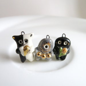 May include: Three ceramic charms, each with a different design. The first charm is black and white with a gold accent, the second is grey with gold accents, and the third is black with a green and gold accent.