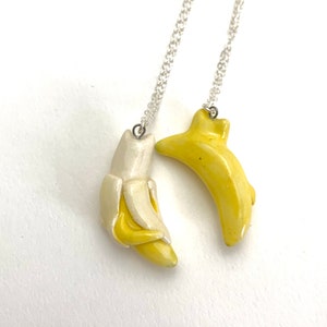 May include: Two silver chain necklaces with banana pendants. One banana pendant is yellow and the other is white.