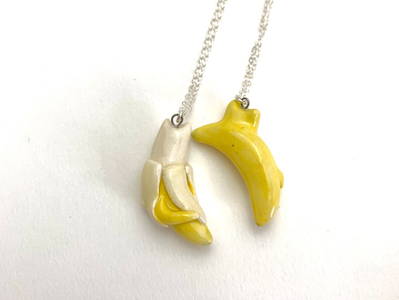 May include: Two silver chain necklaces with banana pendants. One banana pendant is yellow and the other is white.