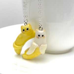 May include: Two ceramic cat necklaces, one yellow and one white, are hanging from silver chains. The cats are shaped like bananas and have painted eyes and mouths.