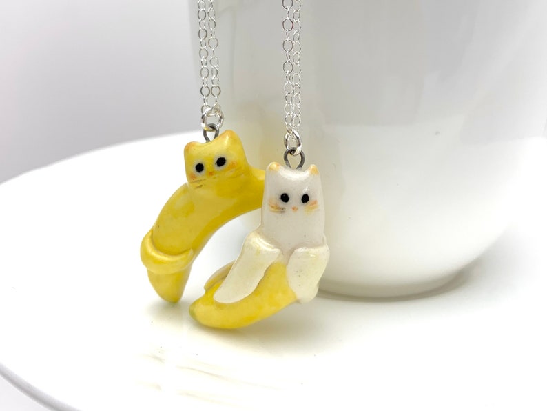 May include: Two ceramic cat necklaces, one yellow and one white, are hanging from silver chains. The cats are shaped like bananas and have painted eyes and mouths.
