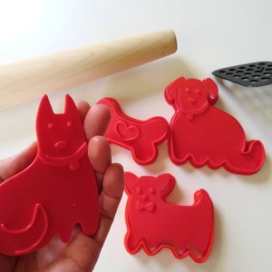 May include: Set of four red plastic dog-shaped cookie cutters. The cutters include a bone, a small dog, a large dog, and a dog with a collar.