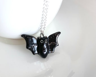May include: A black ceramic bat pendant necklace with a silver chain. The bat has white eyes and a detailed design.
