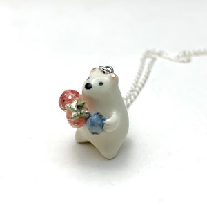 May include: White ceramic bear pendant necklace with a silver chain. The bear is holding a red and green strawberry and a blue blueberry.