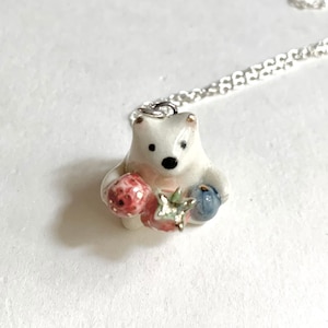 May include: A white ceramic bear pendant necklace with a silver chain. The bear is holding a red strawberry and a blue blueberry.