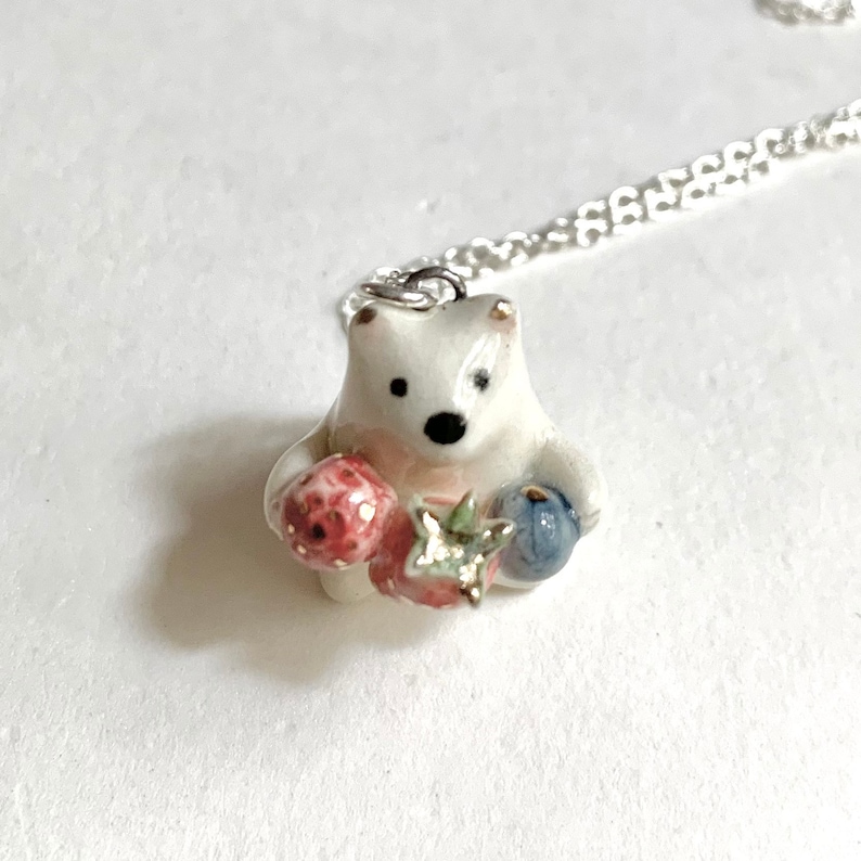 May include: A white ceramic bear pendant necklace with a silver chain. The bear is holding a red strawberry and a blue blueberry.