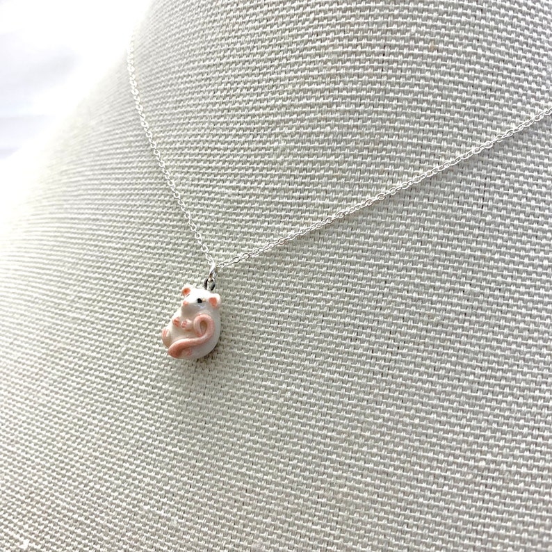 May include: A silver chain necklace with a small white ceramic mouse pendant. The mouse is curled up with its tail wrapped around its body.