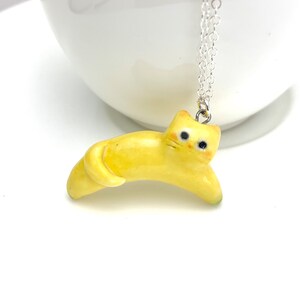 May include: A yellow ceramic cat charm necklace. The cat is lying down with its tail curled around its body. The necklace is made of silver chain.