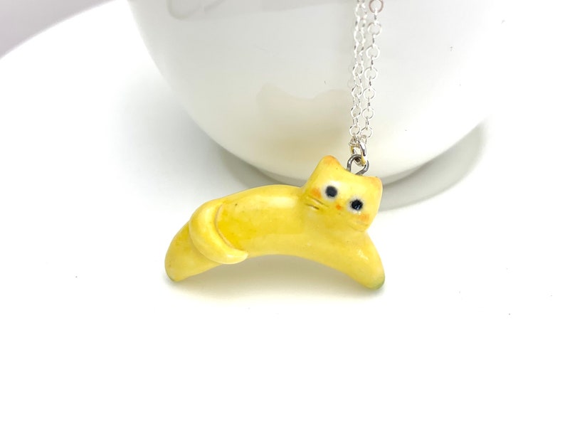 May include: A yellow ceramic cat charm necklace. The cat is lying down with its tail curled around its body. The necklace is made of silver chain.