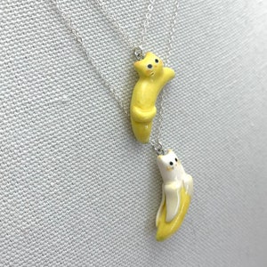 May include: Two silver necklaces with ceramic cat pendants. One cat is yellow and the other is white. Both cats are curled up in a banana shape.