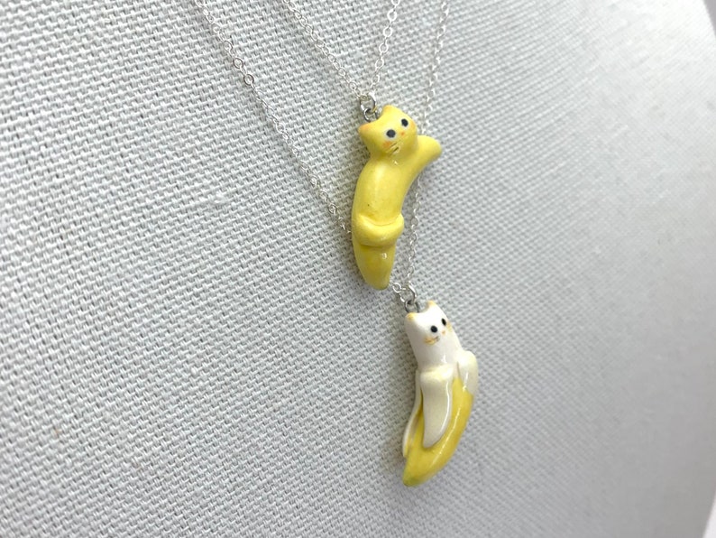 May include: Two silver necklaces with ceramic cat pendants. One cat is yellow and the other is white. Both cats are curled up in a banana shape.