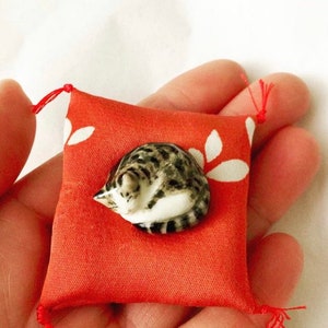 May include: A small, hand-held orange fabric cushion featuring a white leaf pattern and red corner tassels. Resting on the center of the cushion is a miniature ceramic figurine of a curled-up, grey and white tabby cat with dark stripes.