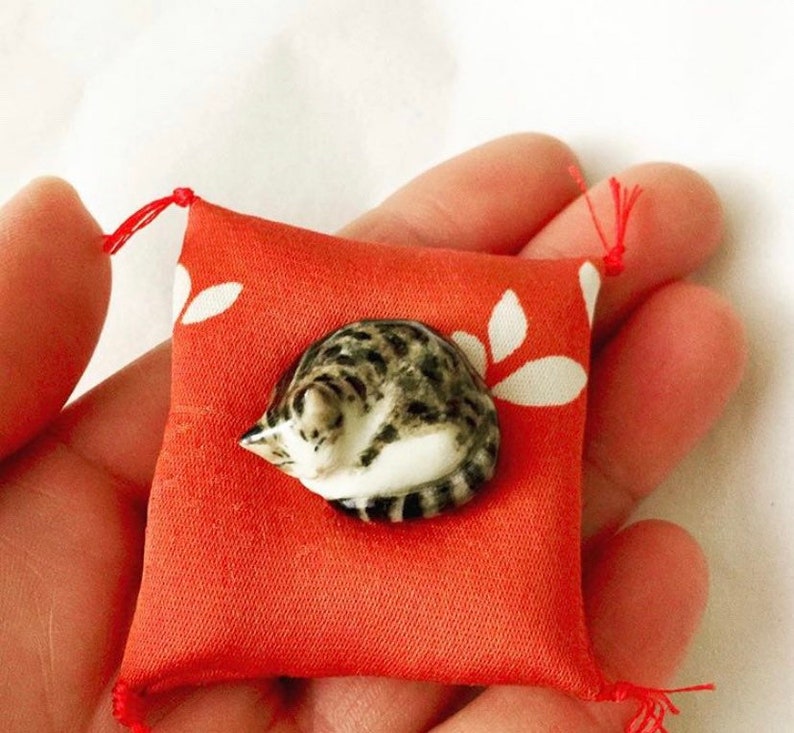 May include: A small, hand-held orange fabric cushion featuring a white leaf pattern and red corner tassels. Resting on the center of the cushion is a miniature ceramic figurine of a curled-up, grey and white tabby cat with dark stripes.
