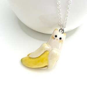 May include: A white ceramic cat figurine wearing a yellow banana costume. The cat has black eyes and a silver chain necklace.