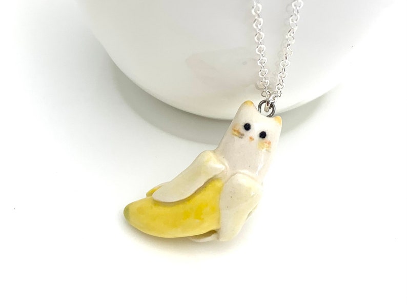 May include: A white ceramic cat figurine wearing a yellow banana costume. The cat has black eyes and a silver chain necklace.