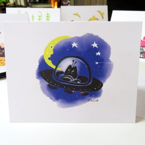 May include: A black cat with green eyes sits in a flying saucer against a purple, blue, and black starry night sky with a yellow crescent moon. The text "Chris Craft" is in the bottom right corner.
