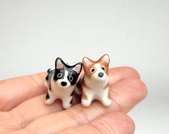 May include: Two miniature ceramic dog figurines, one black, white, and tan, and the other brown and white. Both small animal sculptures feature pointed ears, dark eyes, and a glossy finish, standing side by side in a detailed, hand-painted style.