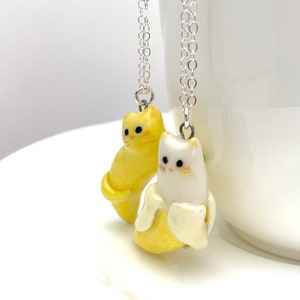 May include: Two ceramic cat necklaces, one yellow and one white, each holding a yellow banana. The necklaces are on silver chains.