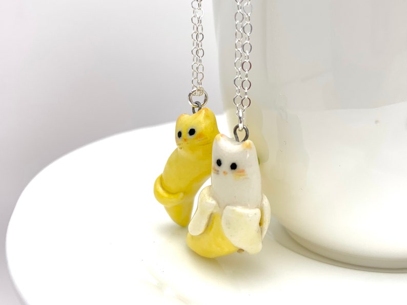 May include: Two ceramic cat necklaces, one yellow and one white, each holding a yellow banana. The necklaces are on silver chains.