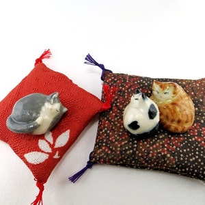May include: Two small square fabric cushions with corner tassels. One cushion is red with a white leaf pattern and a gray ceramic cat figurine. The other is a dark brown patterned cushion with two small ceramic cat figurines, one white and black.