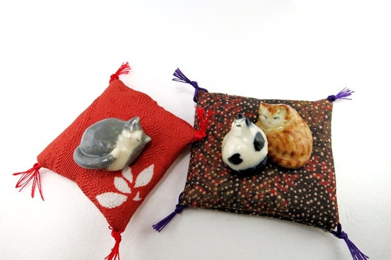 May include: Two small square fabric cushions with corner tassels. One cushion is red with a white leaf pattern and a gray ceramic cat figurine. The other is a dark brown patterned cushion with two small ceramic cat figurines, one white and black.