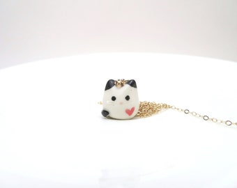 Puede incluir: Un collar con un pequeño colgante de cerámica blanca en forma de gato, con orejas negras, ojos negros y un corazón rosa pintado en el pecho. El colgante está unido a una fina cadena de metal dorado que se extiende hacia la derecha.