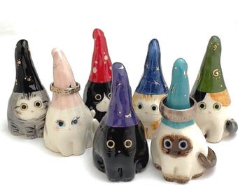 May include: A collection of seven ceramic cat figurines, each wearing a tall, pointed hat. The cats feature various coat patterns and colors, including black, white, grey, and calico. Two of the figurines are wearing silver-toned metal rings.