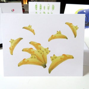 May include: A white card featuring a watercolour illustration of yellow bananas with cat faces. The bananas are arranged in a cluster with some overlapping. The card is blank and ready for a message.
