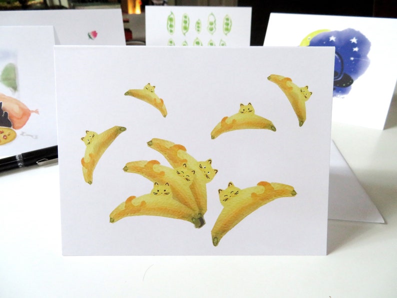 May include: A white card featuring a watercolour illustration of yellow bananas with cat faces. The bananas are arranged in a cluster with some overlapping. The card is blank and ready for a message.