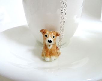 Puede incluir: Un collar con un colgante de cerámica en forma de perro marrón y blanco en una cadena de plata.